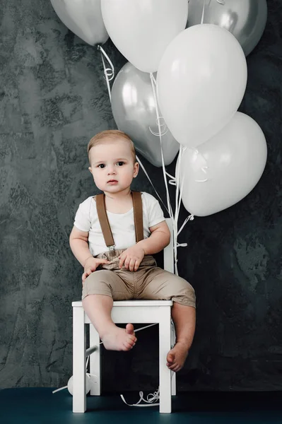 Retrato del adorable niño feliz sonriendo celebrando 1 año de cumpleaños. Un pequeño caballero europeo de un año sentado en una silla y jugando con globos — Foto de Stock