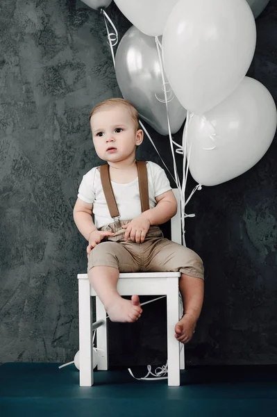 Retrato de adorável menino pequeno feliz sorrindo celebrando 1 ano de aniversário. Um ano de idade europeu pequeno cavalheiro sentado em uma cadeira e brincando com balões — Fotografia de Stock