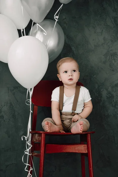 Retrato del adorable niño feliz sonriendo celebrando 1 año de cumpleaños. Un pequeño caballero europeo de un año sentado en una silla y jugando con globos — Foto de Stock