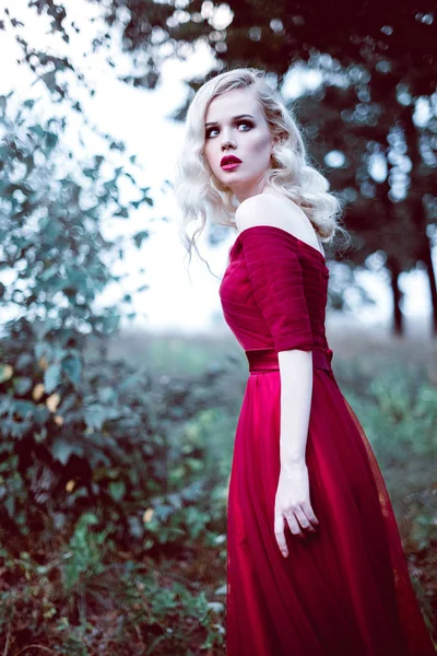 Moda splendida giovane donna bionda in bellissimo vestito rosso in una foresta da favola. atmosfera magica. Ritocco tonificante shot — Foto Stock