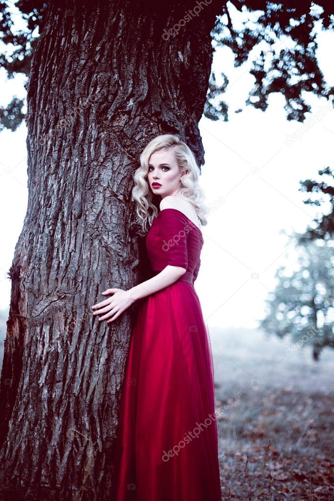Fashion gorgeous young blonde woman in beautiful red dress in a fairy-tale forest. magic atmosphere. Retouched toning shot
