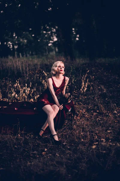 Belle et élégante femme blonde aux lèvres rouges et aux vagues de cheveux portant une nuisette rouge vin posant sur le lit en plein air en automne, style vintage rétro et mode . — Photo