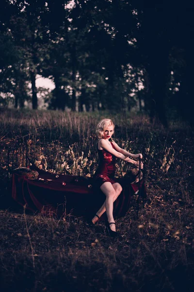 Belle et élégante femme blonde aux lèvres rouges et aux vagues de cheveux portant une nuisette rouge vin posant sur le lit en plein air en automne, style vintage rétro et mode . — Photo