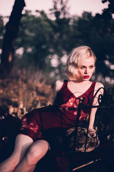Мбаппе и элегантная блондинка с рыжими губами и волнами волос в винно-красном вечернем галстуке, позирующая на кровати под открытым небом осенью, ретро-винтажный стиль и мода . — стоковое фото