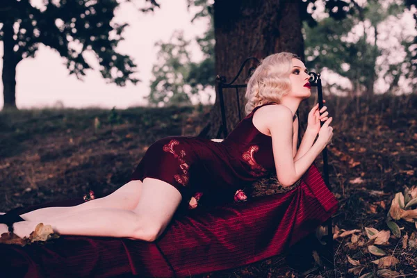 Belle et élégante femme blonde aux lèvres rouges et aux vagues de cheveux portant une nuisette rouge vin posant sur le lit en plein air en automne, style vintage rétro et mode . — Photo