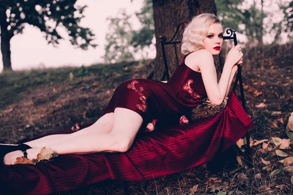 Mujer rubia hermosa y elegante con labios rojos y olas de pelo usando camisón rojo vino posando en la cama al aire libre otoño, estilo vintage retro y moda . — Foto de Stock