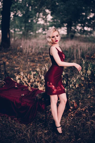 Красива і елегантна блондинка з червоними губами і хвилями волосся у вині червона сукня позує на ліжко на відкритому повітрі осінь, ретро вінтажний стиль і мода . — стокове фото