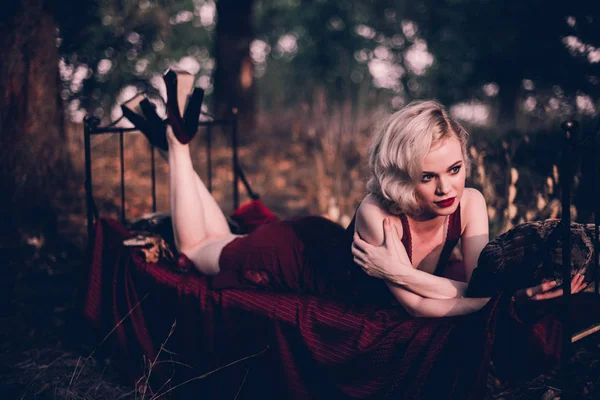 Mooi en elegant blonde vrouw met rode lippen en haren golven dragen wijn rode nachtjapon poseren op het bed buitenshuis herfst, retro vintage stijl en mode. — Stockfoto