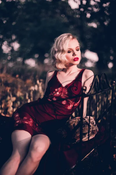 Мбаппе и элегантная блондинка с рыжими губами и волнами волос в винно-красном вечернем галстуке, позирующая на кровати под открытым небом осенью, ретро-винтажный стиль и мода . — стоковое фото