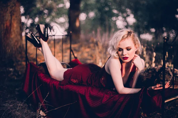 Όμορφο και κομψό ξανθιά γυναίκα με κόκκινα χείλη και τα μαλλιά τα κύματα φορώντας κρασί κόκκινο Νυχτικό ποζάρει στο κρεβάτι σε εξωτερικούς χώρους το φθινόπωρο, ρετρό vintage στυλ και τη μόδα. — Φωτογραφία Αρχείου