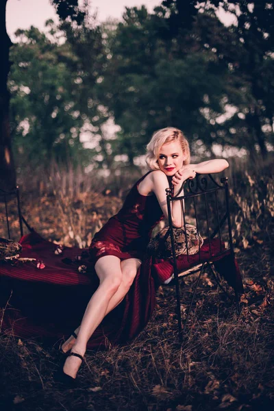 Mooi en elegant blonde vrouw met rode lippen en haren golven dragen wijn rode nachtjapon poseren op het bed buitenshuis herfst, retro vintage stijl en mode. — Stockfoto
