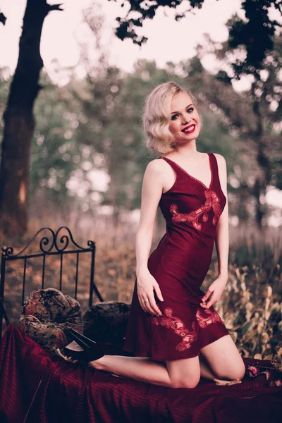 Mujer rubia hermosa y elegante con labios rojos y olas de pelo usando camisón rojo vino posando en la cama al aire libre otoño, estilo vintage retro y moda . — Foto de Stock