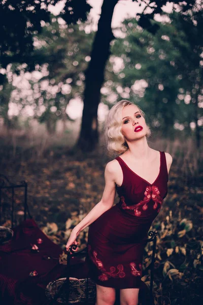 Schöne und elegante blonde Frau mit roten Lippen und Haaren Wellen tragen weinroten Nachthemd posiert auf dem Bett im Freien Herbst, Retro-Vintage-Stil und Mode. — Stockfoto