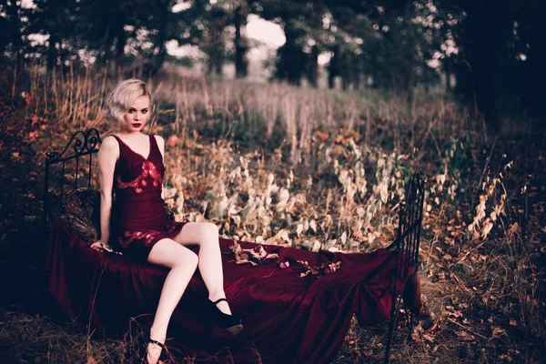 Mooi en elegant blonde vrouw met rode lippen en haren golven dragen wijn rode nachtjapon poseren op het bed buitenshuis herfst, retro vintage stijl en mode. — Stockfoto