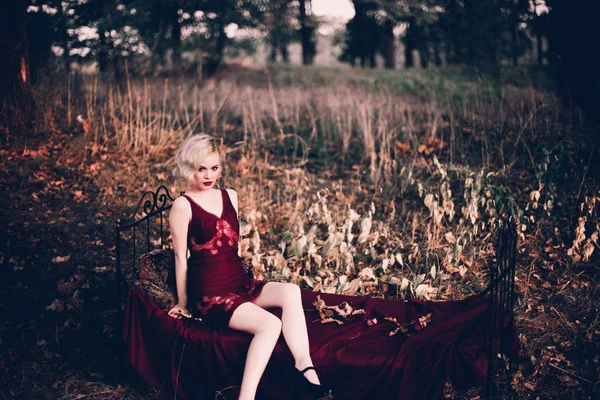 Belle et élégante femme blonde aux lèvres rouges et aux vagues de cheveux portant une nuisette rouge vin posant sur le lit en plein air en automne, style vintage rétro et mode . — Photo
