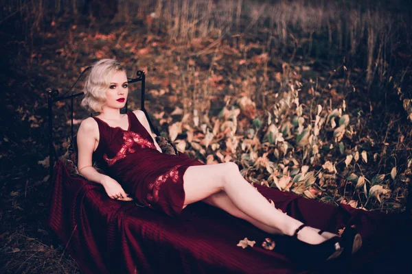 Schöne und elegante blonde Frau mit roten Lippen und Haaren Wellen tragen weinroten Nachthemd posiert auf dem Bett im Freien Herbst, Retro-Vintage-Stil und Mode. — Stockfoto