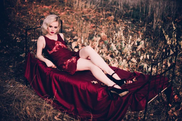 Schöne und elegante blonde Frau mit roten Lippen und Haaren Wellen tragen weinroten Nachthemd posiert auf dem Bett im Freien Herbst, Retro-Vintage-Stil und Mode. — Stockfoto