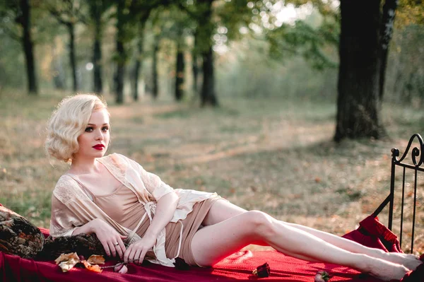 Piękna i elegancka blond kobieta z czerwonymi ustami i włosów fale noszenia beżowy nightie, pozowanie na łóżku na zewnątrz, jesień, retro styl vintage i mody. sztuki, tonowanie strzał — Zdjęcie stockowe