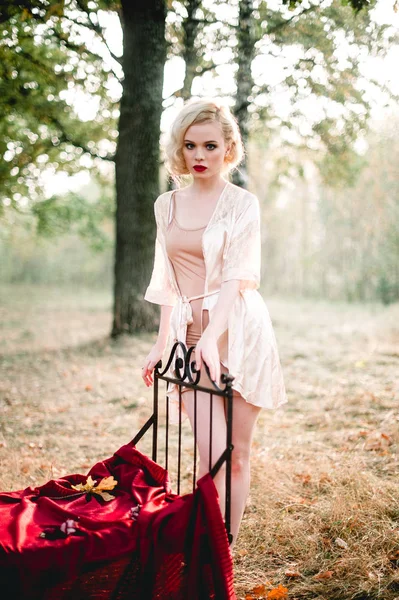 Krásná a elegantní blondýnka s červenými rty a vlasy vlny nosí béžové noční košilku pózuje na posteli venku podzim, retro vintage styl a módy. výtvarného umění tónování shot — Stock fotografie