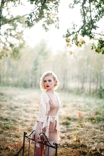 Красива і елегантна блондинка з червоними губами і хвилями волосся носить бежеву сукню, позуючи на ліжку на відкритому повітрі осінь, ретро вінтажний стиль і моду. тонкий художній тонізуючий знімок — стокове фото