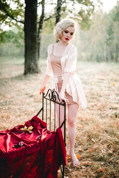 Bella ed elegante donna bionda con labbra rosse e onde di capelli che indossa camicia da notte beige in posa sul letto all'aperto autunno, stile vintage retrò e moda. colpo tonificante arte fine — Foto Stock