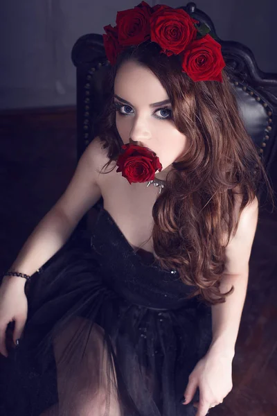 Belle jeune femme avec des fleurs de roses dans les cheveux, portant une robe noire avec maquillage sur fond sombre, atmosphère gothique. rouge foncé lips.close-up mode retouchée portrait — Photo