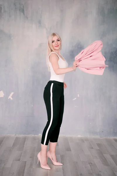 Aantrekkelijke positieve middelbare leeftijd blonde vrouw draagt roze jas en broek met broek strepen met een mooie glimlach tegen een teruglopende muur rechtstreeks kijken naar de camera poseren. Mode geretoucheerd p — Stockfoto