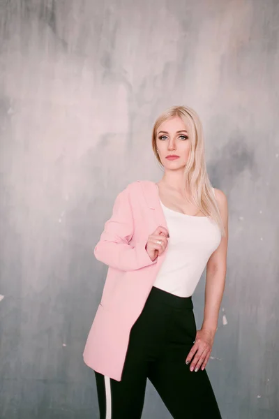 Attractive positive middle-aged blond woman wearing pink jacket and pants with trouser stripes with a beautiful smile posing against a receding wall looking directly at the camera. Fashion retouched p — Stock Photo, Image