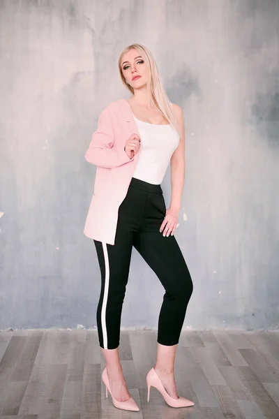 Attractive positive middle-aged blond woman wearing pink jacket and pants with trouser stripes with a beautiful smile posing against a receding wall looking directly at the camera. Fashion retouched p — Stock Photo, Image