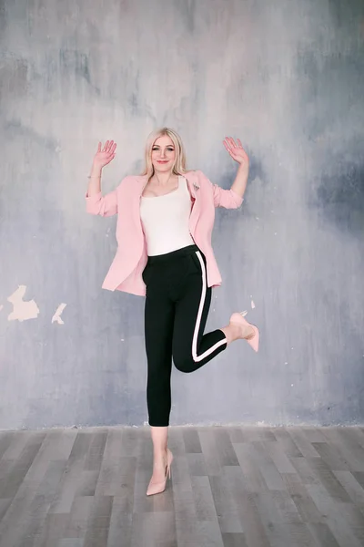 Attractive positive middle-aged blond woman wearing pink jacket and pants with trouser stripes with a beautiful smile posing against a receding wall looking directly at the camera. Fashion retouched p