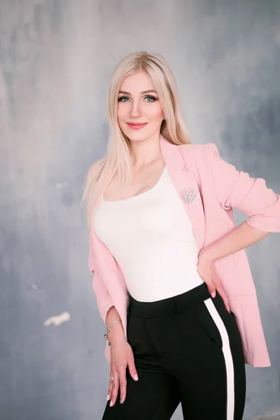 Attractive positive middle-aged blond woman wearing pink jacket and pants with trouser stripes with a beautiful smile posing against a receding wall looking directly at the camera. Fashion retouched p — Stock Photo, Image