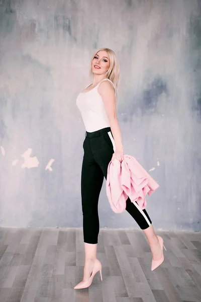Attractive positive middle-aged blond woman wearing pink jacket and pants with trouser stripes with a beautiful smile posing against a receding wall looking directly at the camera. Fashion retouched p — Stock Photo, Image