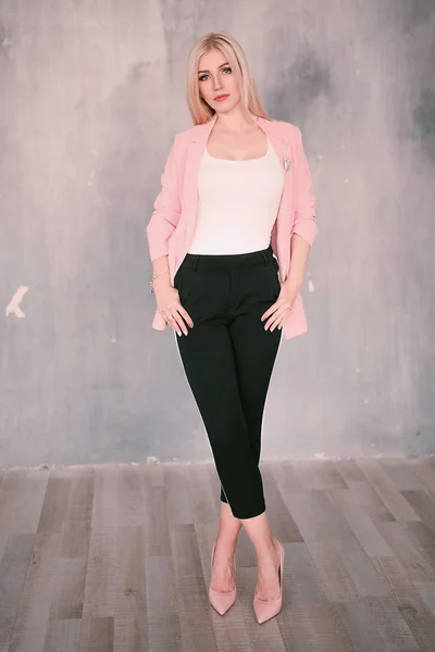 Attractive positive middle-aged blond woman wearing pink jacket and pants with trouser stripes with a beautiful smile posing against a receding wall looking directly at the camera. Fashion retouched p — Stock Photo, Image