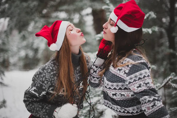 Two young teenage hipster girl friends together.Close up fashion portrait of two sisters hugs and having fun winter time,wearing red santa hats and sweater,best friends couple outdoors, snowy weather — Stock Photo, Image