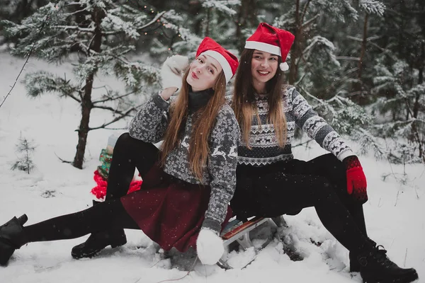 两个年轻的少女时髦女孩朋友在一起。关闭了的两个姐妹拥抱时尚肖像和乐趣冬天的时候，红色的圣诞帽，身穿毛衣，最好的朋友夫妇在户外，下雪的天气 — 图库照片