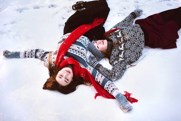 Två unga tonåriga hipster flicka vänner grupp. Nära håll mode porträtt av två systrar kramar och ha roligt ligga på snö i vinter tid skog, bära tröjor och halsdukar, bästa vänner par utomhus — Stockfoto