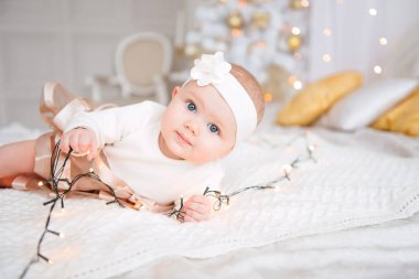 Baby girl wearing cute dress and headband, lies on a white cover in festively decorated room with garland of lights. With surprise watches in the camera, on a background a set of bright fires. Warm be clipart
