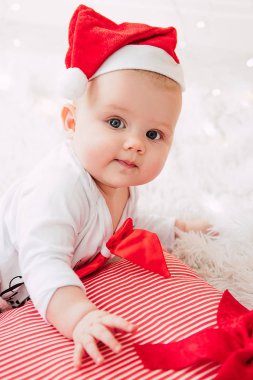 Baby girl wearing cute red dress and santa hat, near christmas tree in festively decorated room with garland of lights. White and red colors of christmas and new year atmosphere. Concept of a happy fa clipart