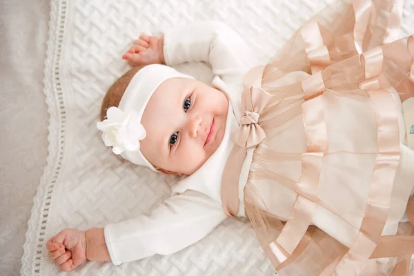 Baby flicka klädd i söt klänning och pannband, ligger på en vit cover i festligt dekorerade rum med krans av lampor. Med förvåning klockor i kameran, på en bakgrund en uppsättning ljusa bränder. Varm vara — Stockfoto