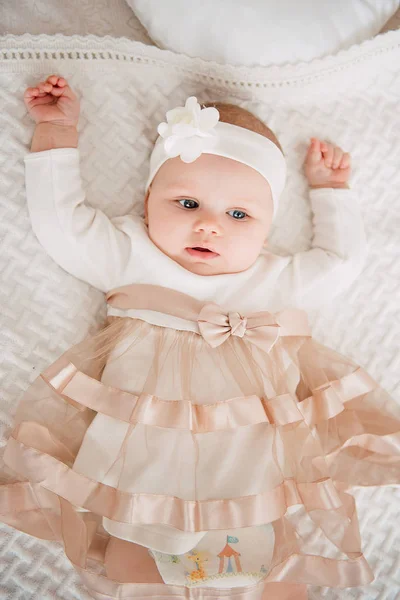 Niña vistiendo lindo vestido y diadema, se encuentra en una cubierta blanca en la habitación festivamente decorada con guirnalda de luces. Con relojes sorpresa en la cámara, sobre un fondo un conjunto de fuegos brillantes. Cálido. — Foto de Stock