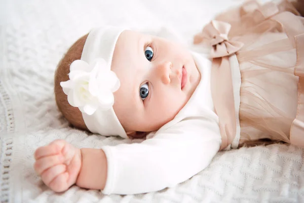 Baby girl wearing cute dress and headband, lies on a white cover in festively decorated room with garland of lights. With surprise watches in the camera, on a background a set of bright fires. Warm be