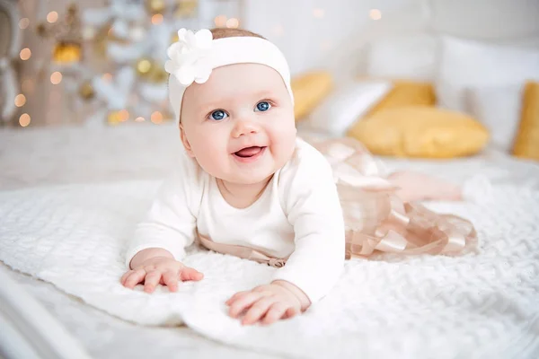 Baby girl wearing cute dress and headband, lies on a white cover in festively decorated room with garland of lights. With surprise watches in the camera, on a background a set of bright fires. Warm be