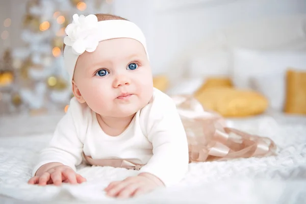 Niña vistiendo lindo vestido y diadema, se encuentra en una cubierta blanca en la habitación festivamente decorada con guirnalda de luces. Con relojes sorpresa en la cámara, sobre un fondo un conjunto de fuegos brillantes. Cálido. — Foto de Stock