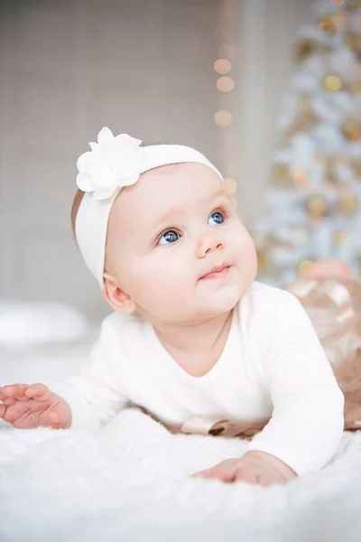 Baby girl wearing cute dress and headband, lies on a white cover in festively decorated room with garland of lights. With surprise watches in the camera, on a background a set of bright fires. Warm be