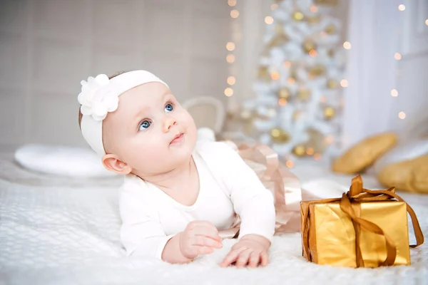 着てかわいい女の赤ちゃんのドレスし、鉢巻き、ガーランド ライトのお祝いに飾られた部屋で白いカバーであります。驚きと明るい火のセットを背景に、カメラの時計します。暖かくします。 — ストック写真