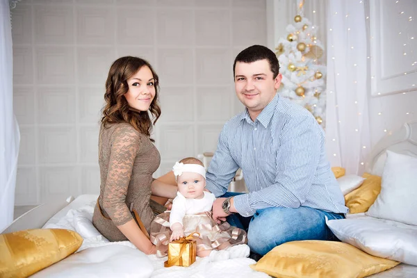 Niña vistiendo lindo vestido y diadema con la madre y el padre, cerca del árbol de Navidad en la habitación festivamente decorada con guirnalda de luces. Los colores cálidos de color beige y oro de la Navidad y el ambiente de año nuevo — Foto de Stock