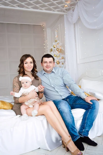 Bebê menina vestindo vestido bonito e headband com mãe e pai, perto da árvore de Natal em quarto festivamente decorado com guirlanda de luzes. Quente bege e ouro cores de Natal e ano novo atmosférico — Fotografia de Stock