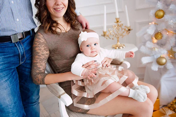 Baby flicka klädd i söt klänning och pannband med mor och far, nära julgran i festligt inrett rum med krans av lampor. Varma beige och guld färger av jul och nyår atmosfär — Stockfoto