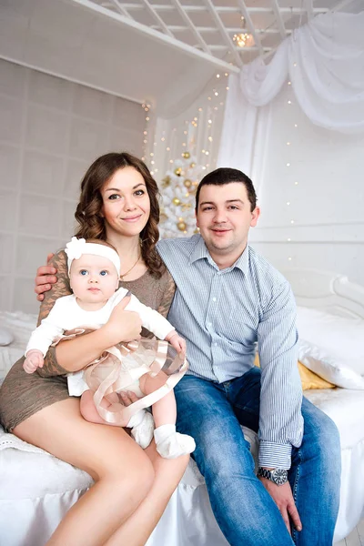 Bambina vestita in modo carino e fascia con madre e padre, vicino all'albero di Natale in camera decorata a festa con ghirlanda di luci. Colori caldi beige e oro di Natale e Capodanno atmospher — Foto Stock