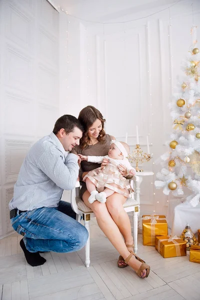 ドレス着てかわいい女の赤ちゃんと母と父のクリスマス ツリーの近くで鉢巻きはお祝いには内装をライトのガーランドで。暖かいクリスマスとお正月の雰囲気のベージュとゴールドの色 — ストック写真